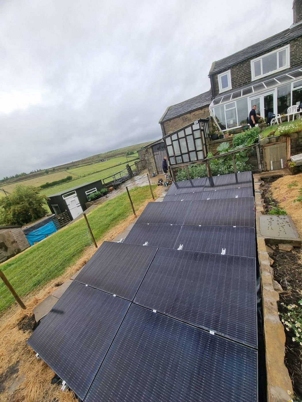 Rows of black solar panels on a ground-mount frame in a sloping garden, with a stone house behind and hills beyond