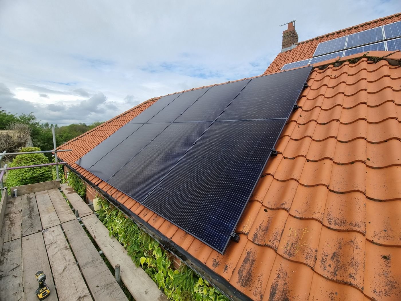 New all-black solar panels on an orange clay pantile roof, with scaffolding and an older solar array higher up