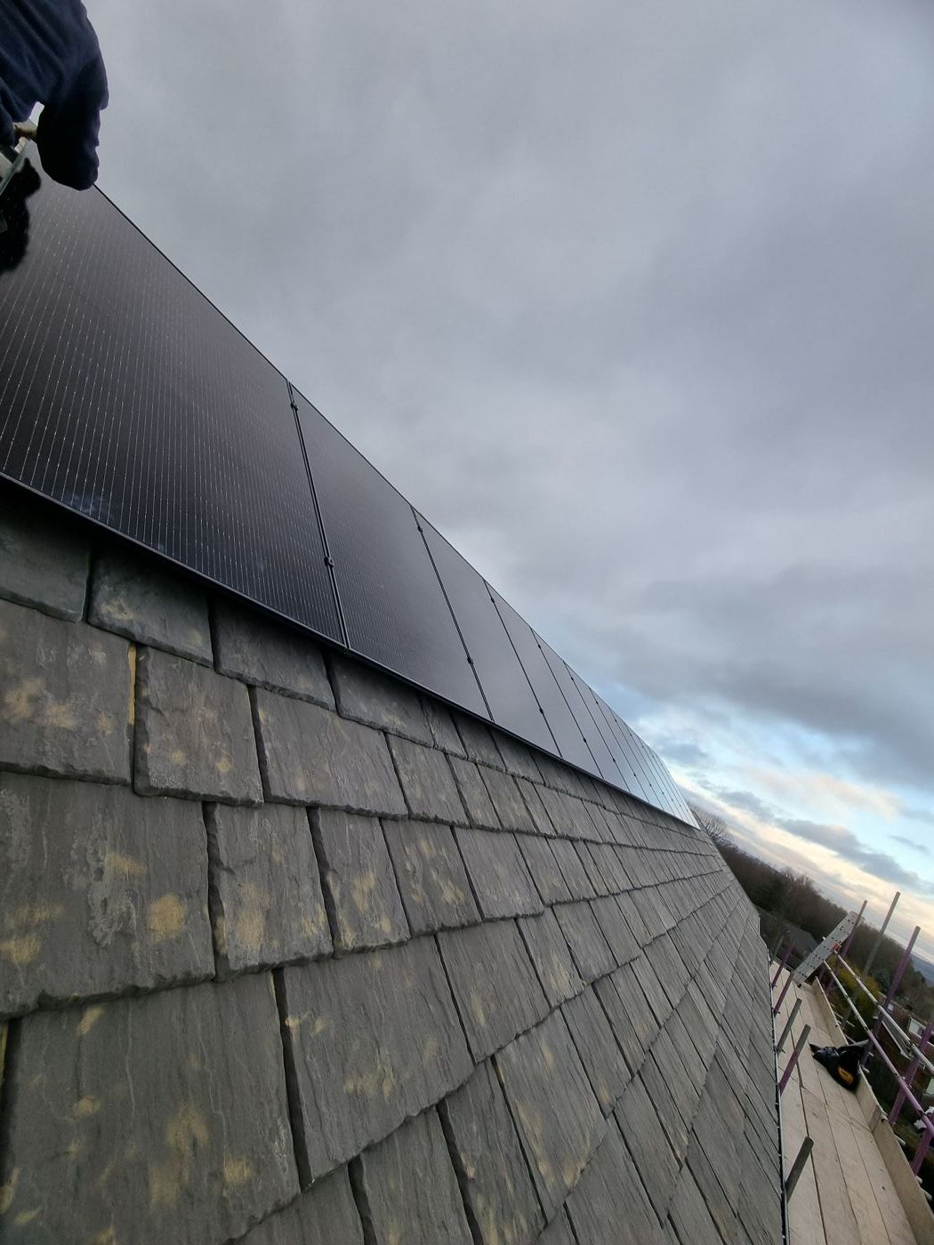 A row of all-black solar panels fitted on a grey stone-slate roof under a cloudy sky